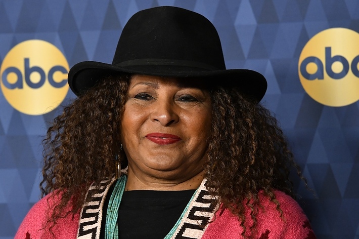 Pam Grier at an event, wearing a black hat, beaded necklace, and a pink-striped blazer