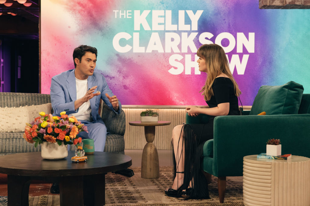 Henry Golding, in a suit, chatting with Kelly Clarkson on her show set
