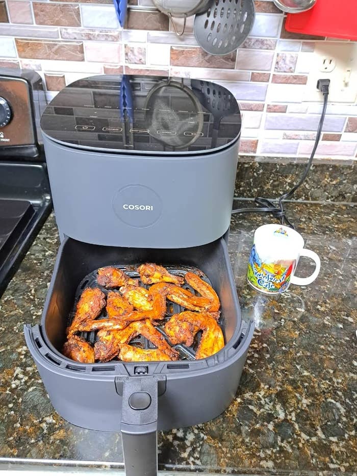 Cosori air fryer on kitchen counter with cooked chicken wings inside, next to a mug