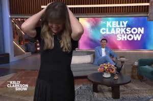 Woman in a black dress stands with her hands in her hair on the set of The Kelly Clarkson Show, host smiling in background