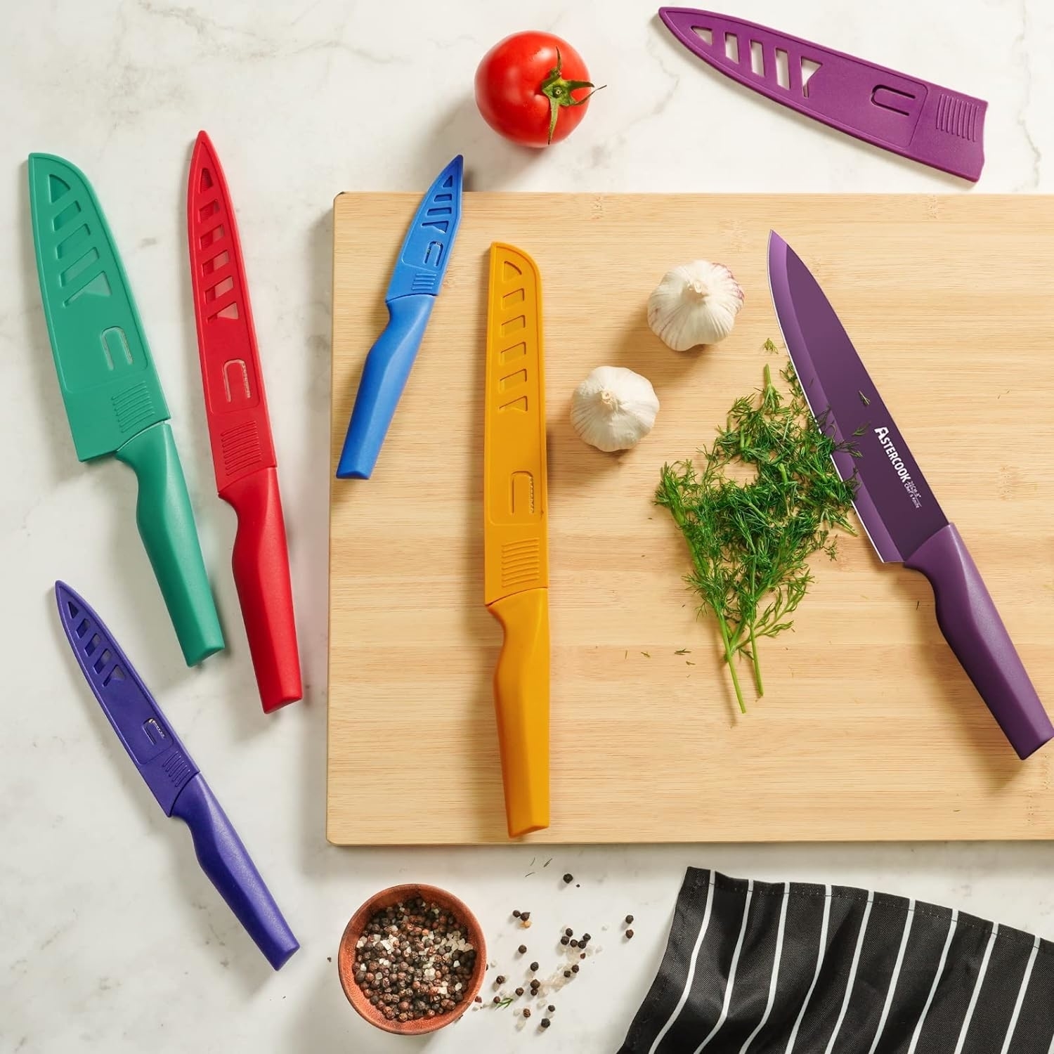 Six kitchen knives with blade guards on a counter next to chopping board with herbs and garlic