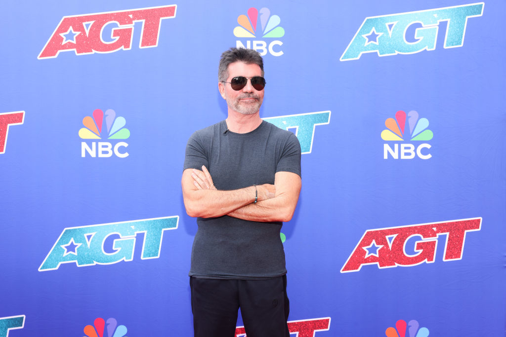 Simon Cowell standing with arms crossed in a casual t-shirt at the AGT backdrop