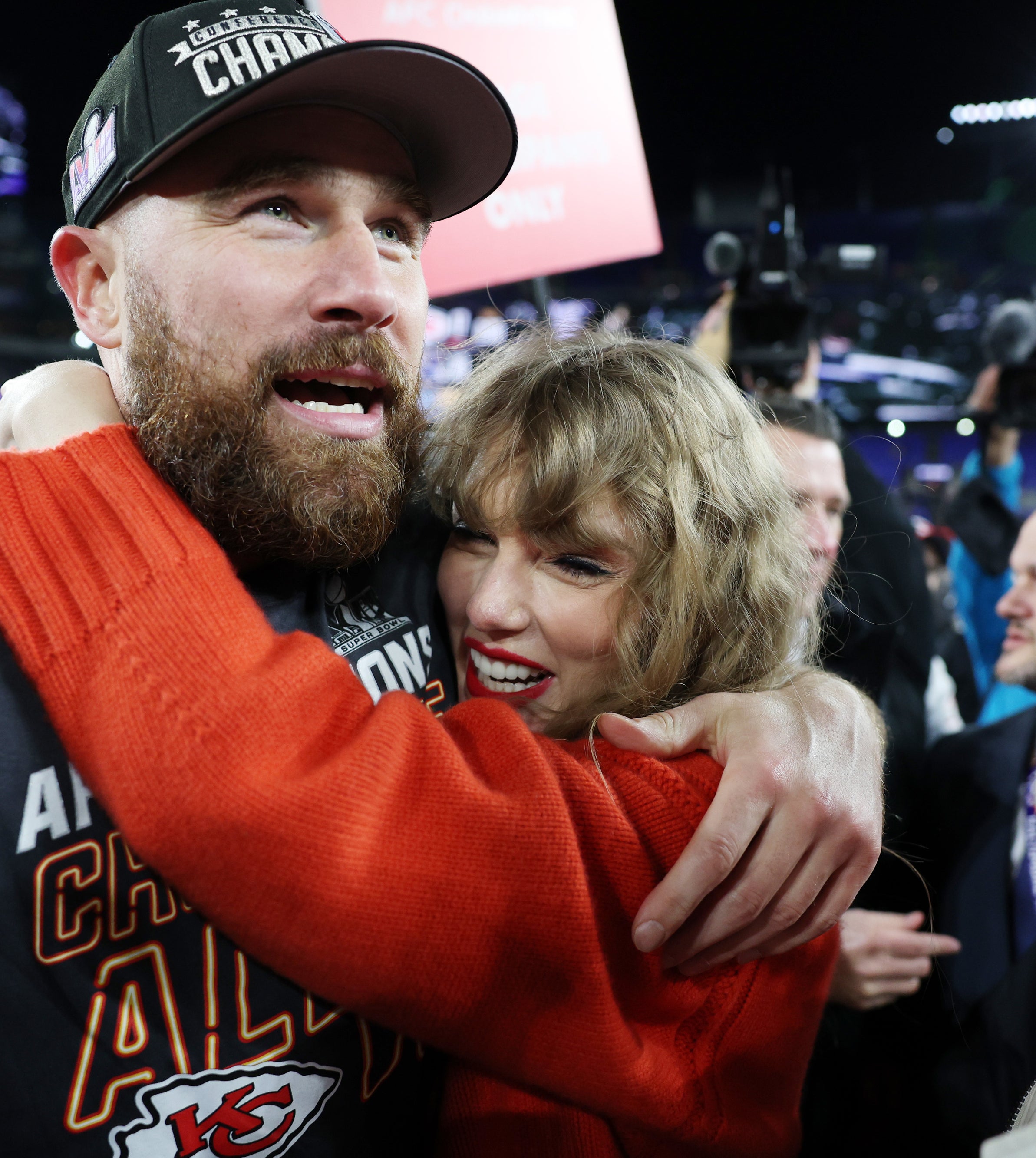 Travis Kelce in a baseball cap hugs a smiling Taylor Swift at an event, surrounded by cameras and onlookers