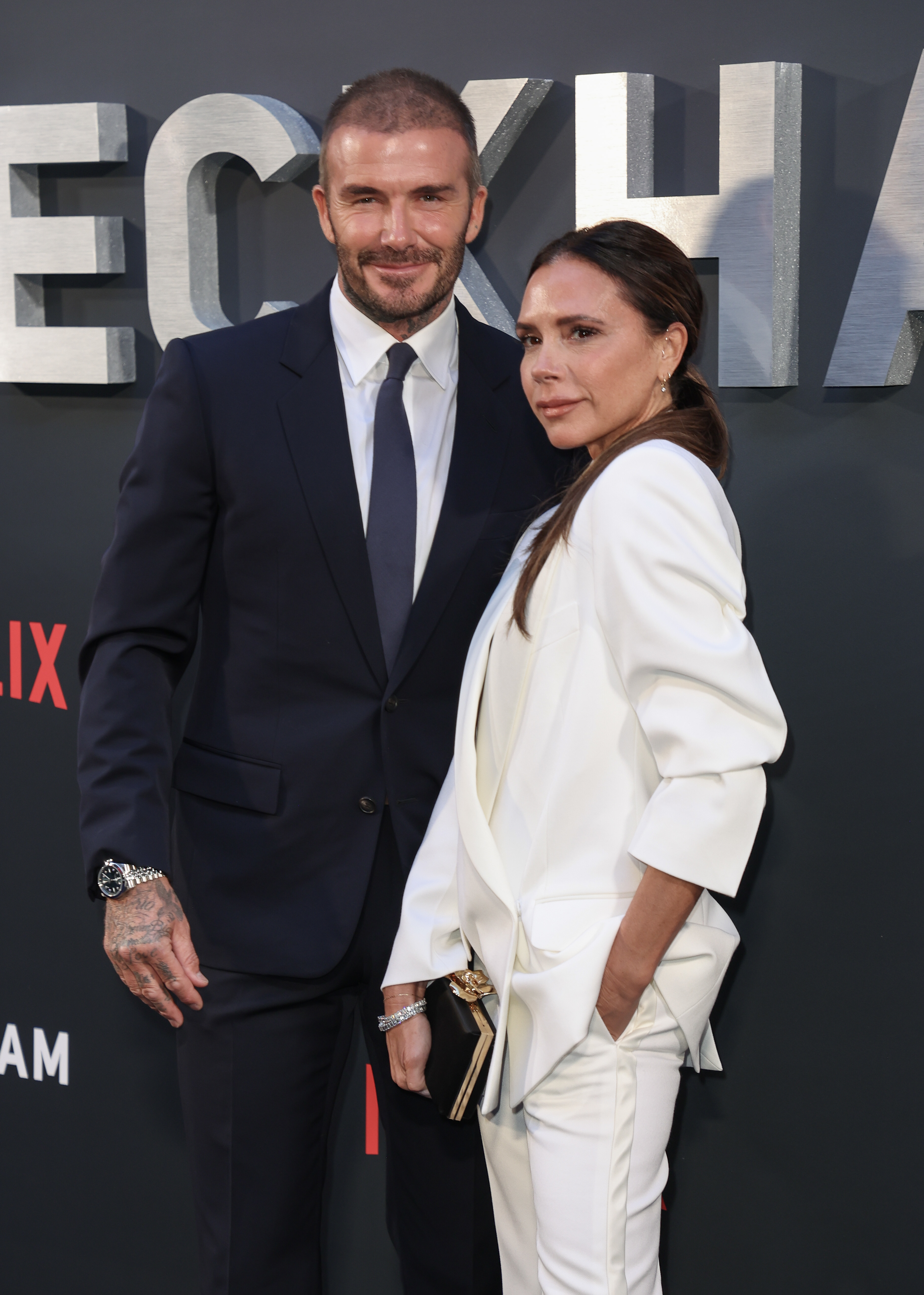 David Beckham and Victoria Beckham pose together at a media event. David is wearing a classic suit and Victoria is wearing a relaxed pantsuit