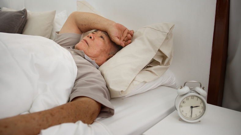 Elderly person lying in bed looking at an alarm clock, appears to be having trouble sleeping