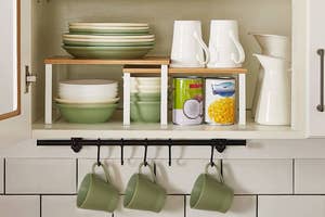 Open shelf in a kitchen with neatly arranged dishes and canned goods, and mugs hanging underneath