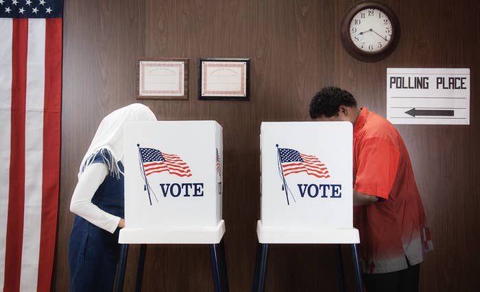 Two individuals at voting booths with &quot;VOTE&quot; banners, American flag in background, indicating a polling place