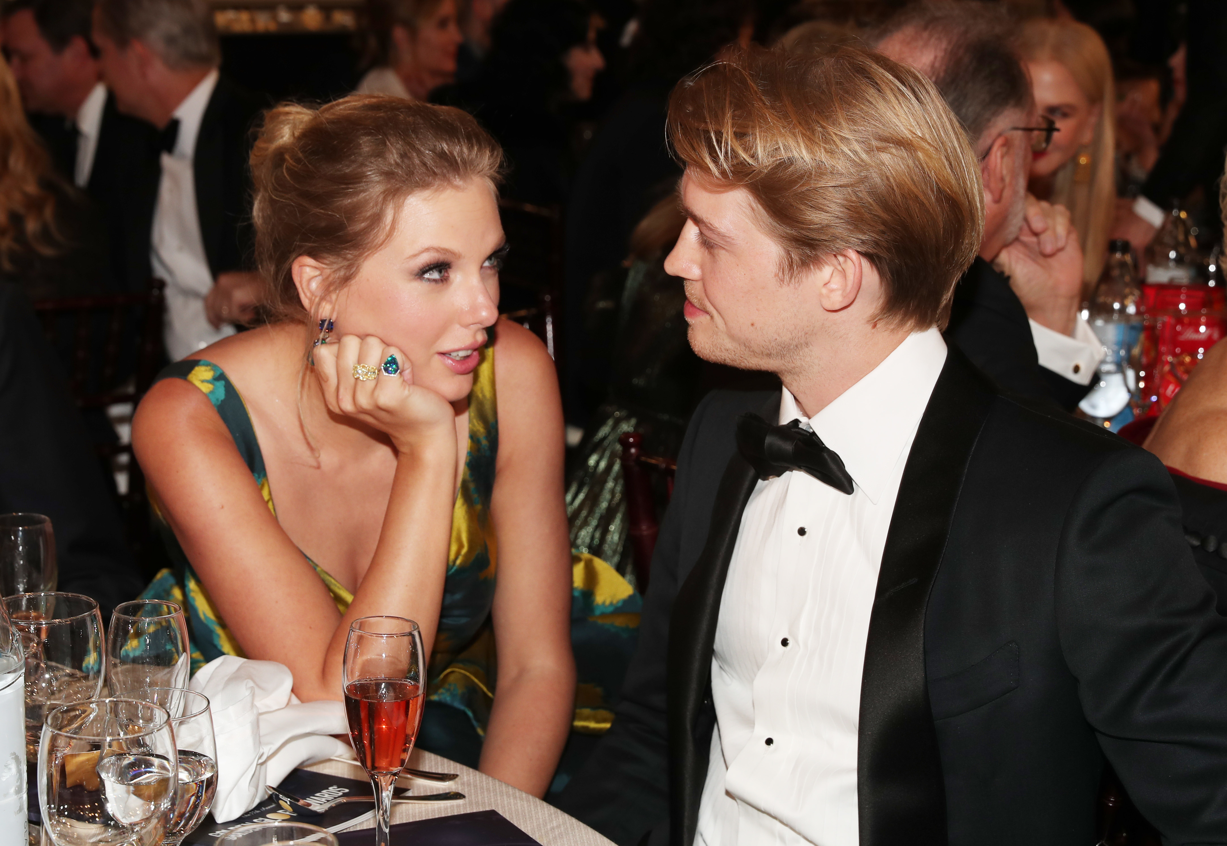 Taylor Swift conversing with Joe Alwyn as they sit at a table at a formal event