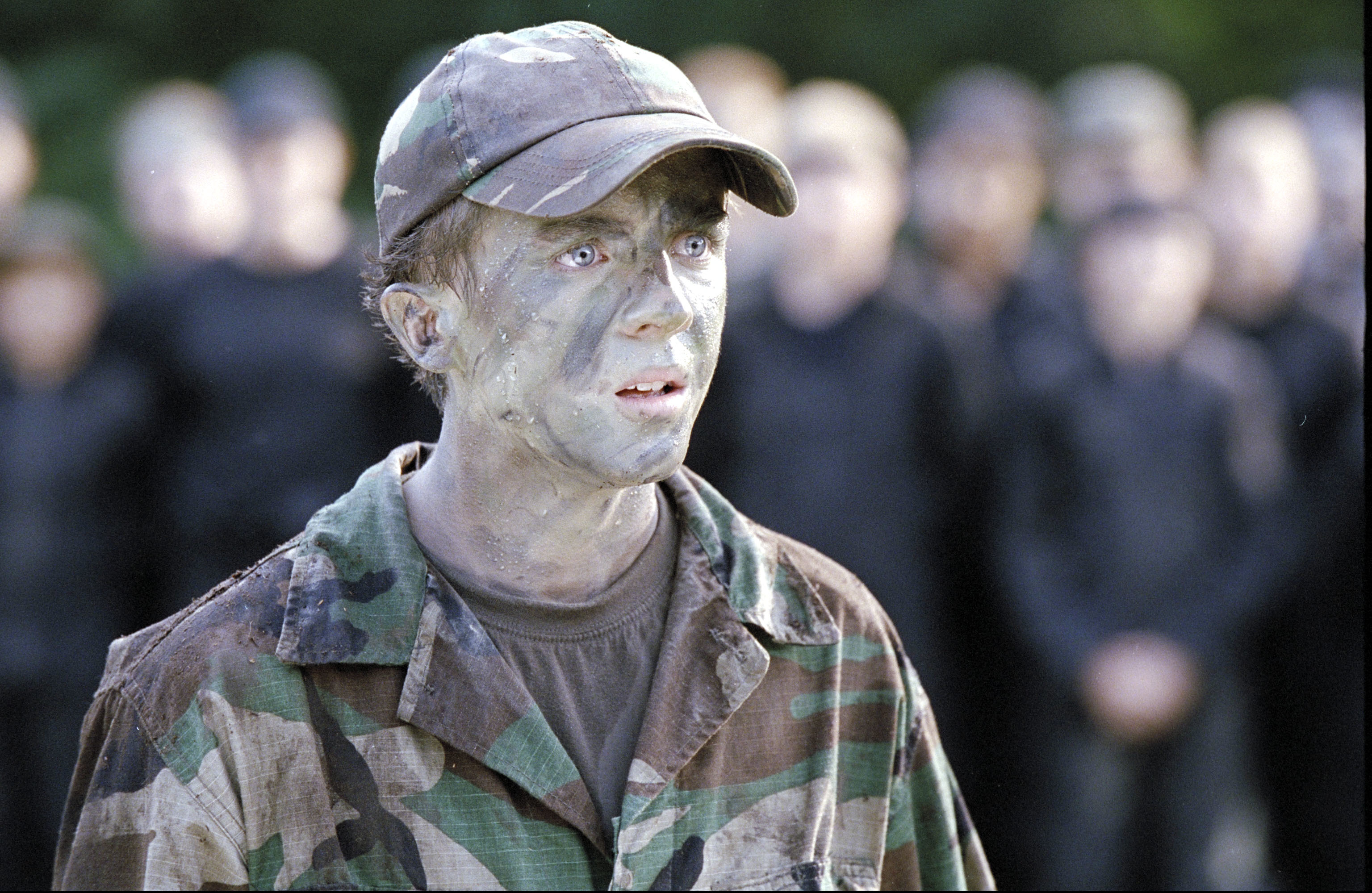 Person in camouflage with face paint, expression of focus, others blurred in background