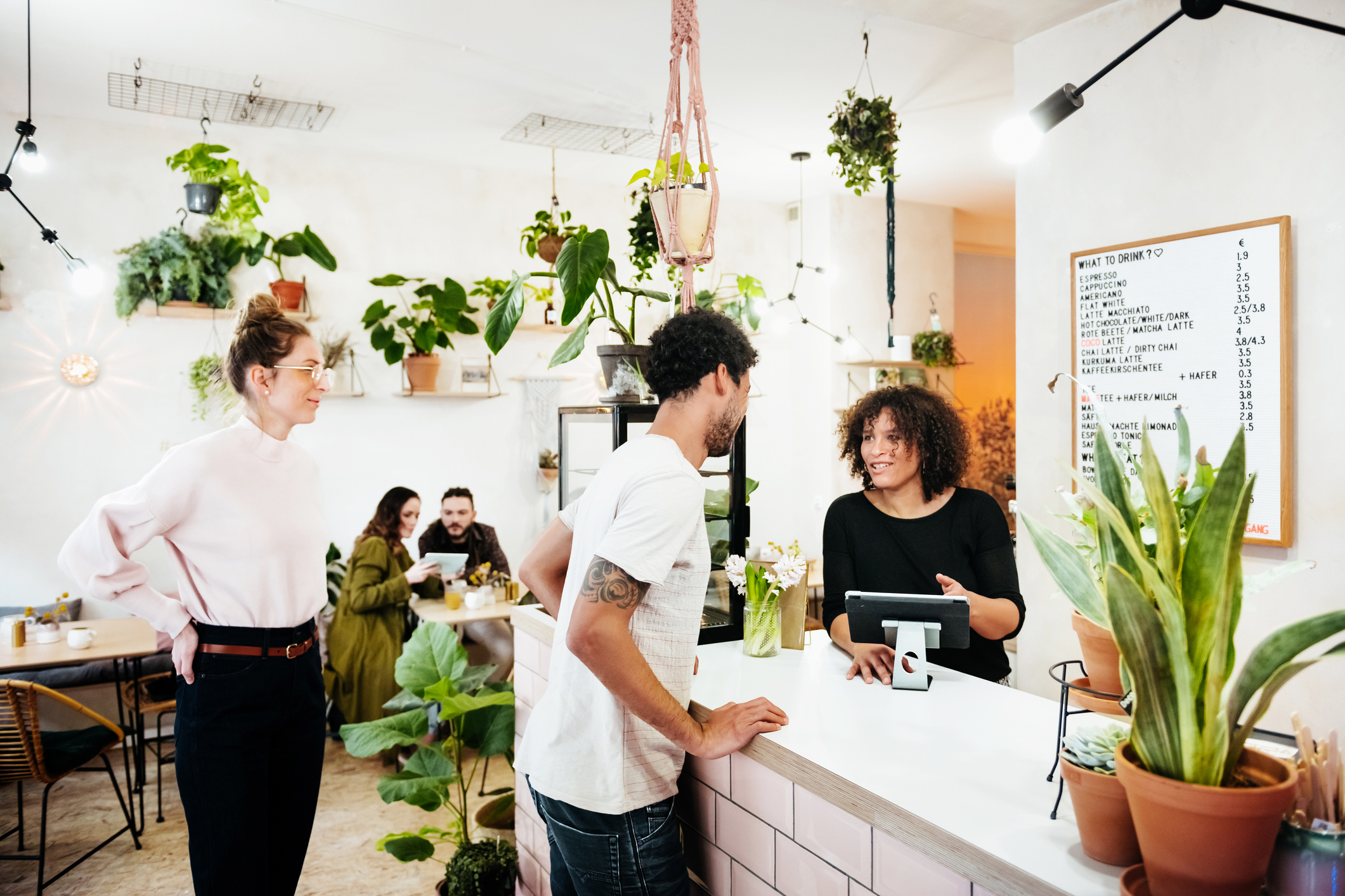 Person ordering at the counter of a cozy cafe with hanging plants and customers in the background. Menu board visible