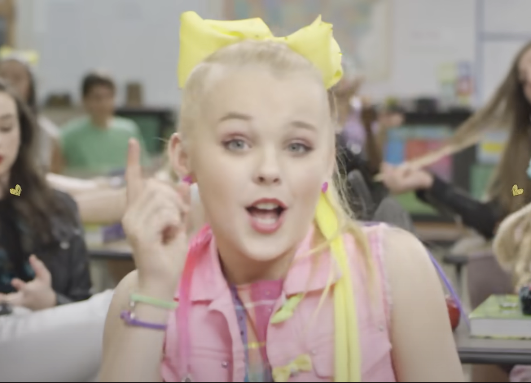 JoJo Siwa in a classroom setting, wearing a signature bow, gesturing with her finger raised