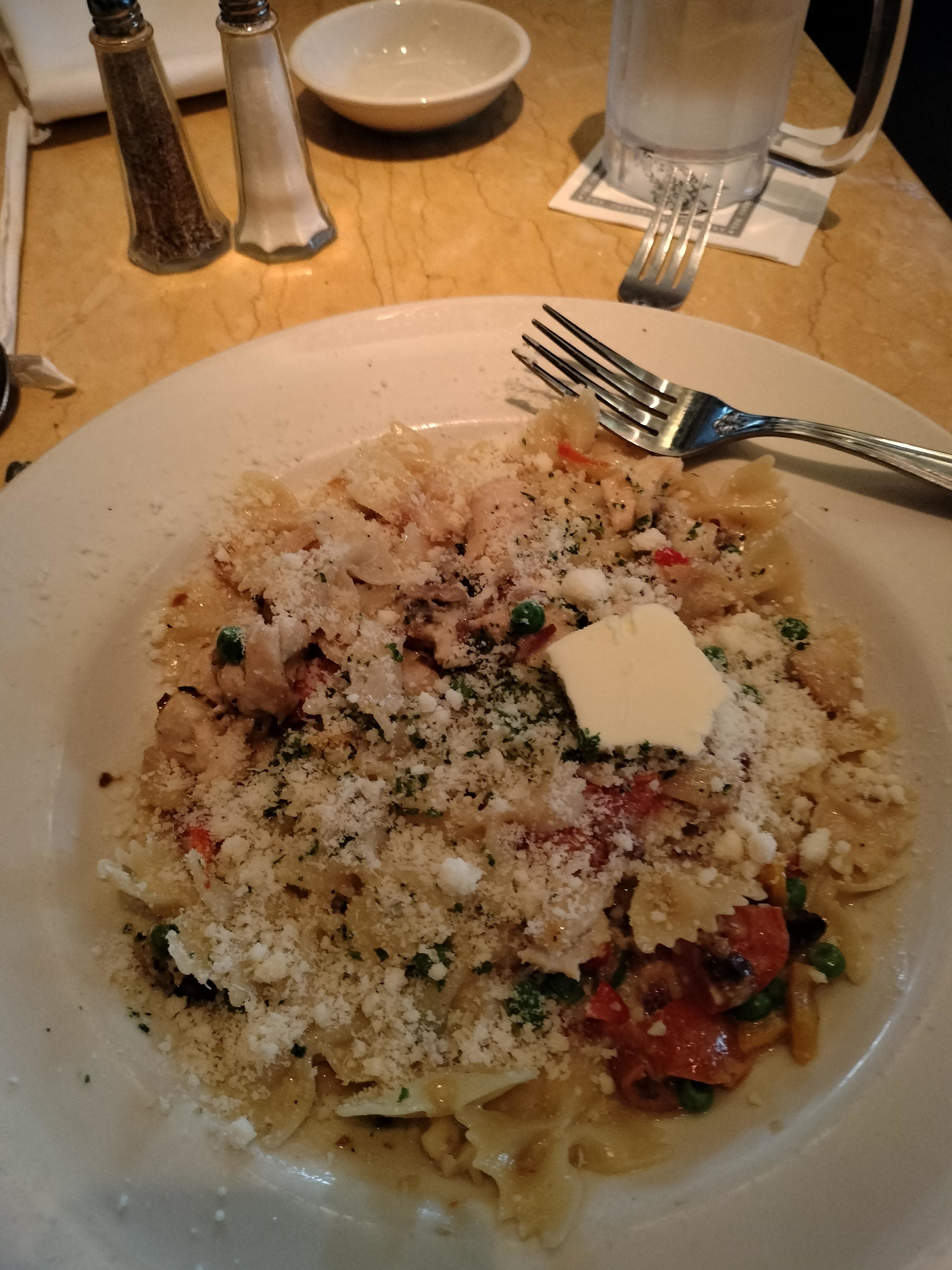 A plate of pasta with chicken, vegetables, and a slice of butter, topped with grated cheese, on a table with salt and pepper shakers