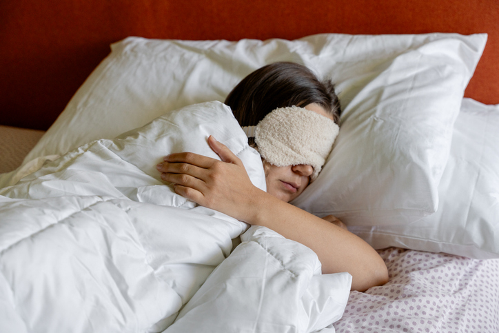 Person sleeping in bed with eye mask, head on pillow, arm peeking out of blanket