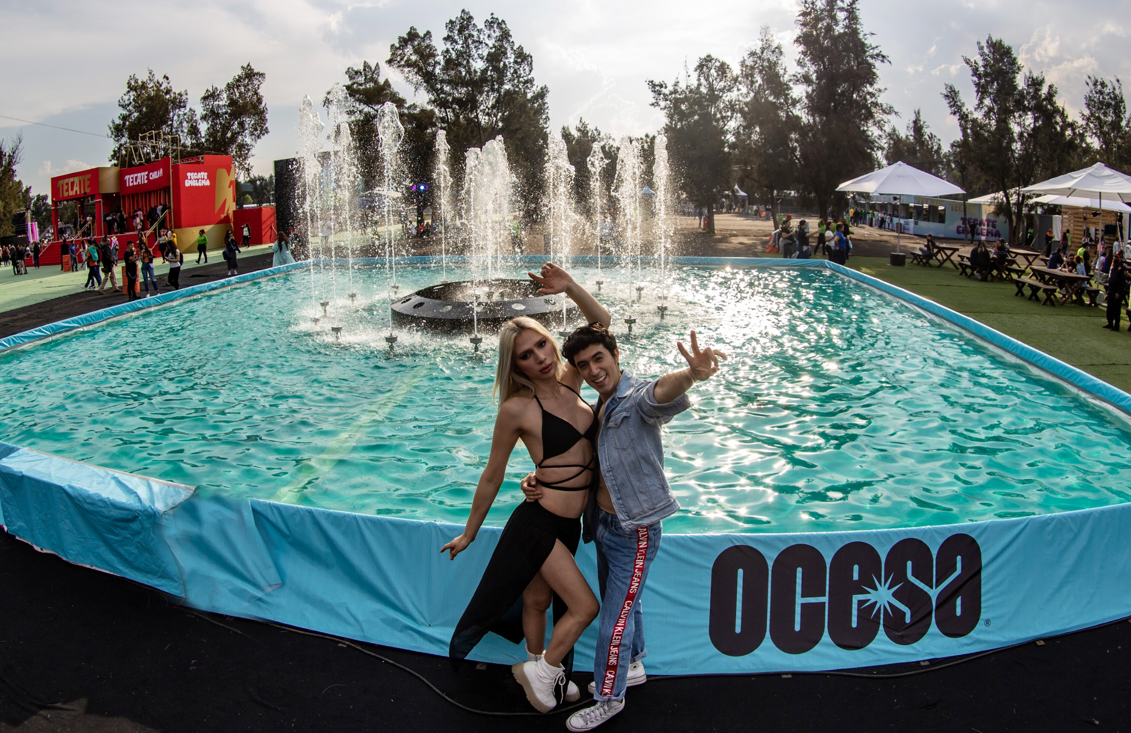 Dos personas posando con los brazos levantados frente a una fuente en un evento de música
