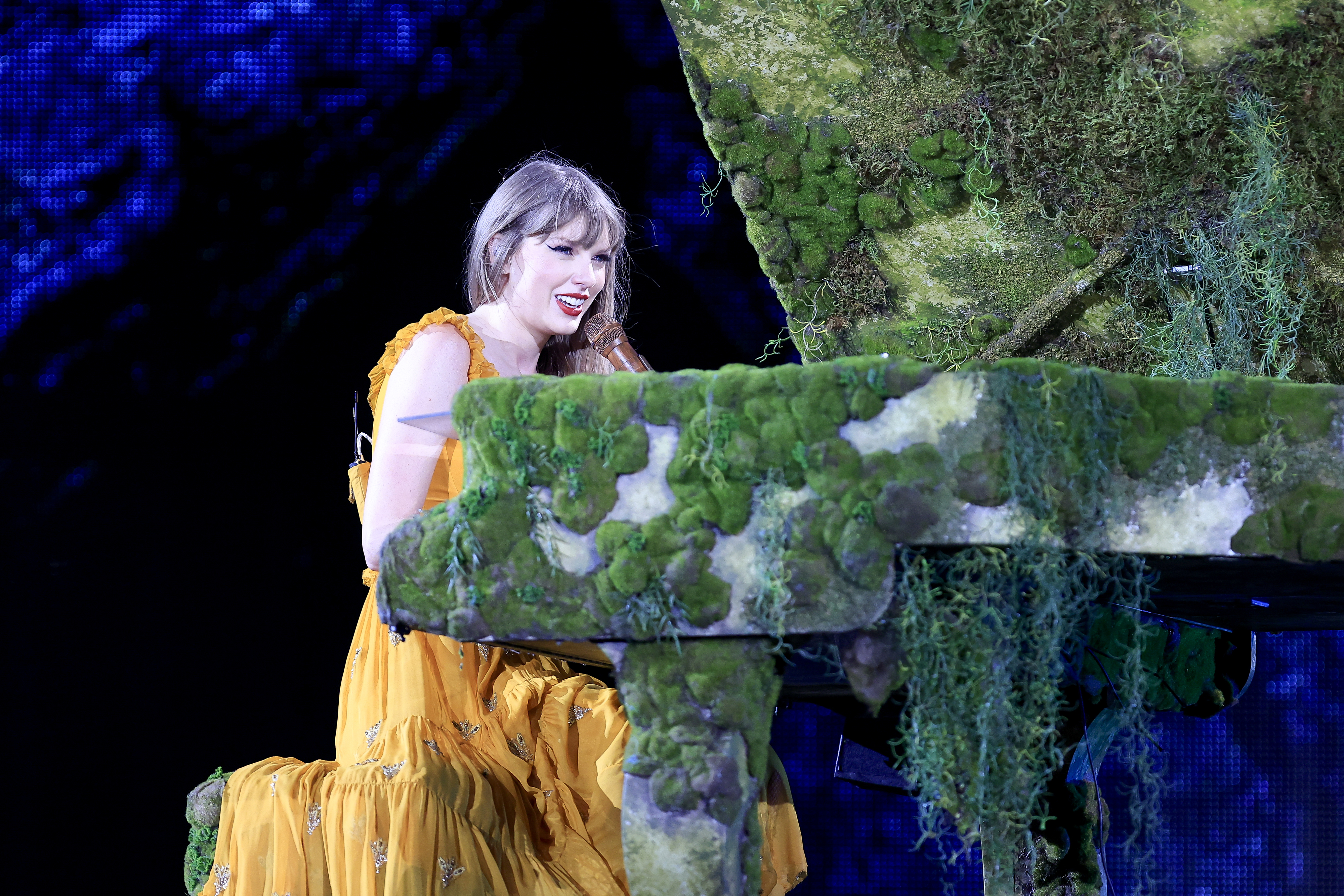 Taylor Swift at piano on stage with mossy decoration in a flowing dress