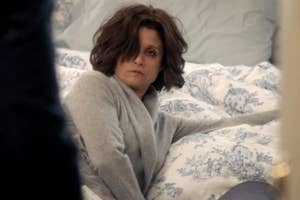 Woman sitting on bed with blue-patterned linens, wearing a grey cardigan and appearing contemplative
