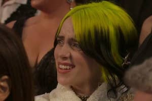 Billie Eilish with vibrant green and black hair, smiling, in a white textured outfit