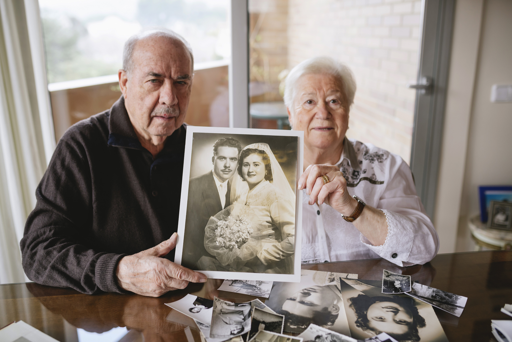 Two elderly individuals holding a framed wedding photo, surrounded by loose photographs on a table