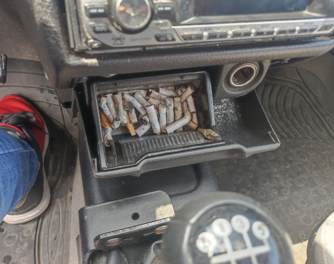 Open ashtray in a car filled with cigarette butts, with a partial view of the dashboard above and gear shift to the right