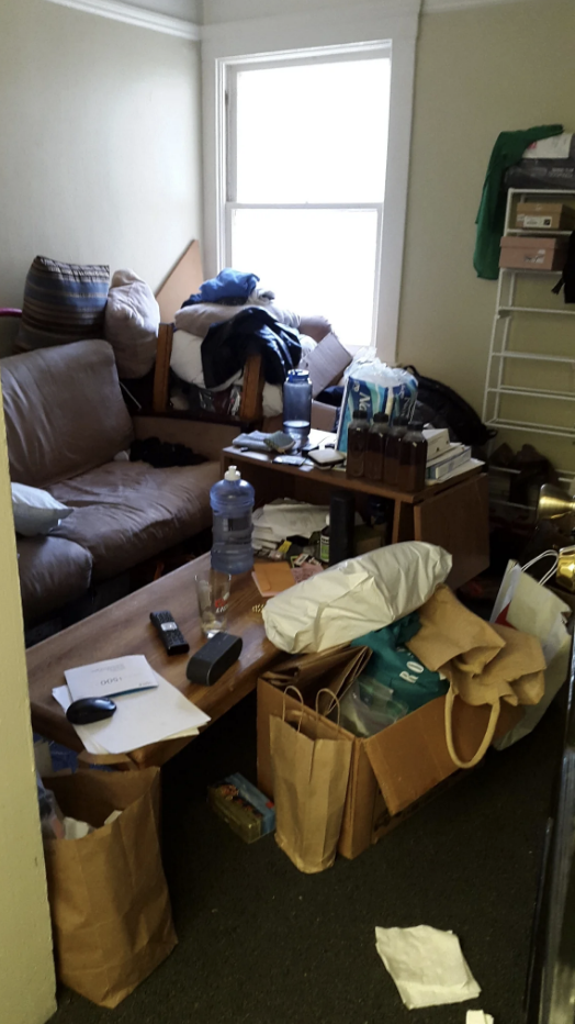 A cluttered room with a sofa, scattered clothing, boxes, and various items on surfaces