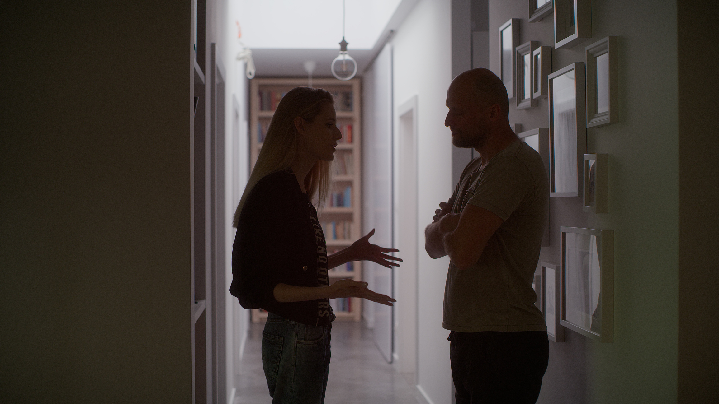 Two people engaged in a conversation in a dimly lit hallway with photo frames on the wall
