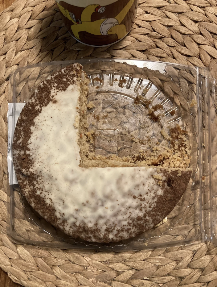 Half-eaten round crumb cake with white icing, in a clear plastic container, on a woven table mat next to a cup