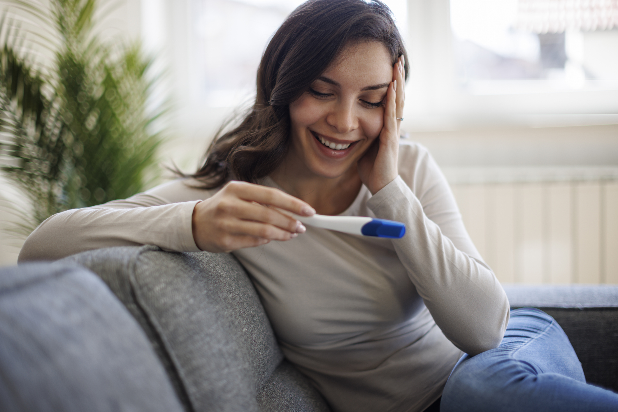 Woman smiling at a positive pregnancy test while sitting on a couch