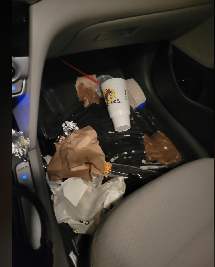 Car floor littered with various trash including a plastic cup, wrappers, and paper bags