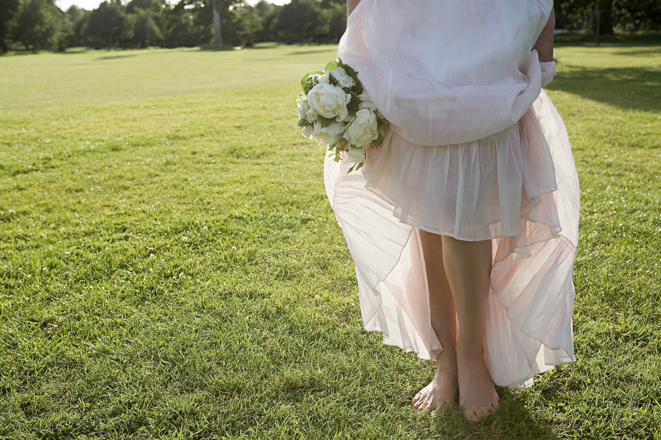 Person in a wedding dress holding a bouquet, standing barefoot on grass
