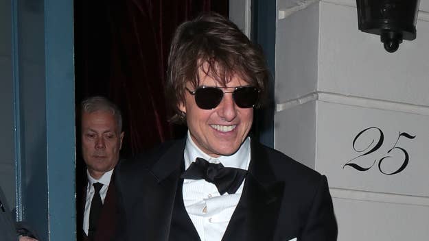 Tom Cruise smiles, wearing a suit and sunglasses at a night event