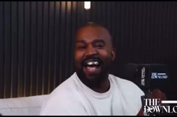Kanye West laughs in a studio setting holding a microphone with "THE DOWNLOAD" text visible