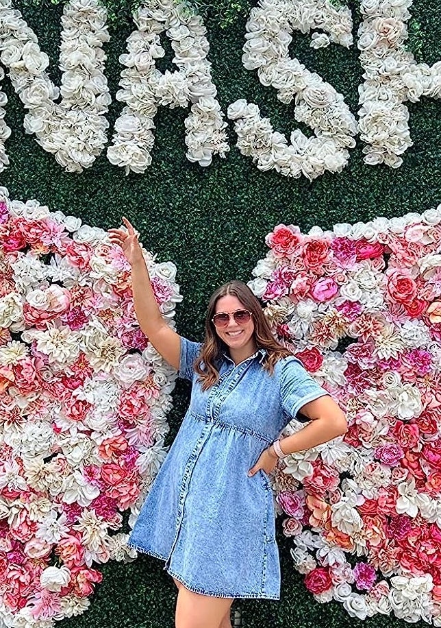Woman posing in front of floral wing display with &quot;NASH&quot; signage, wearing a denim dress and white boots