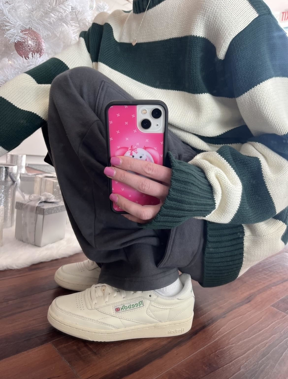 Person sitting with crossed legs, holding phone with a pink case, wearing green and white striped sweater and beige sneakers