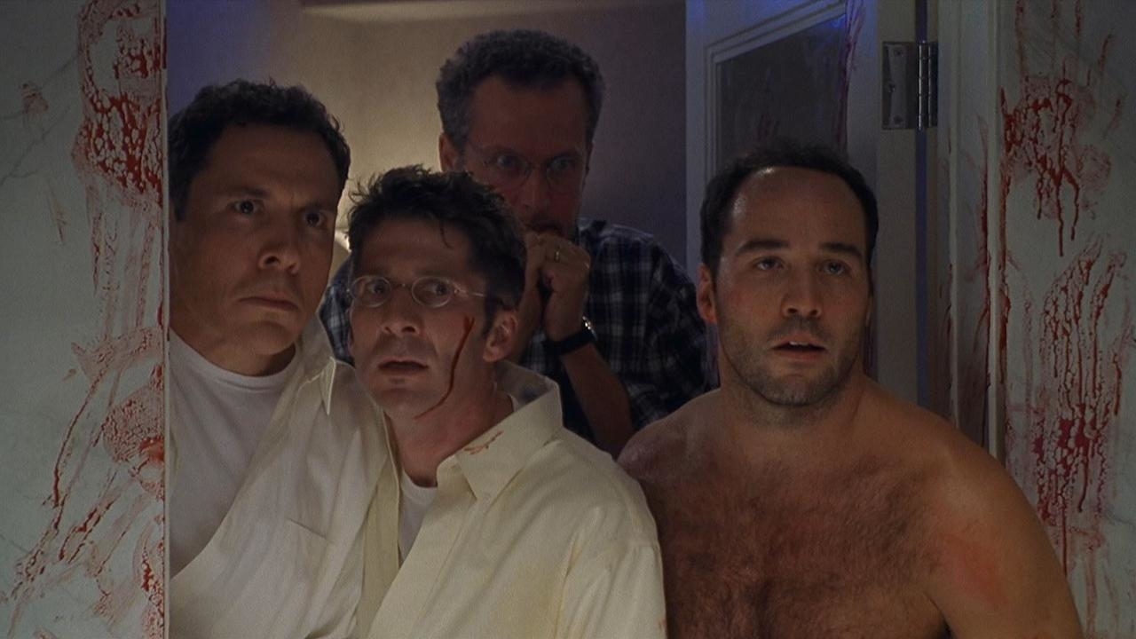 Four men with shocked expressions standing in a room with blood spattered on the wall