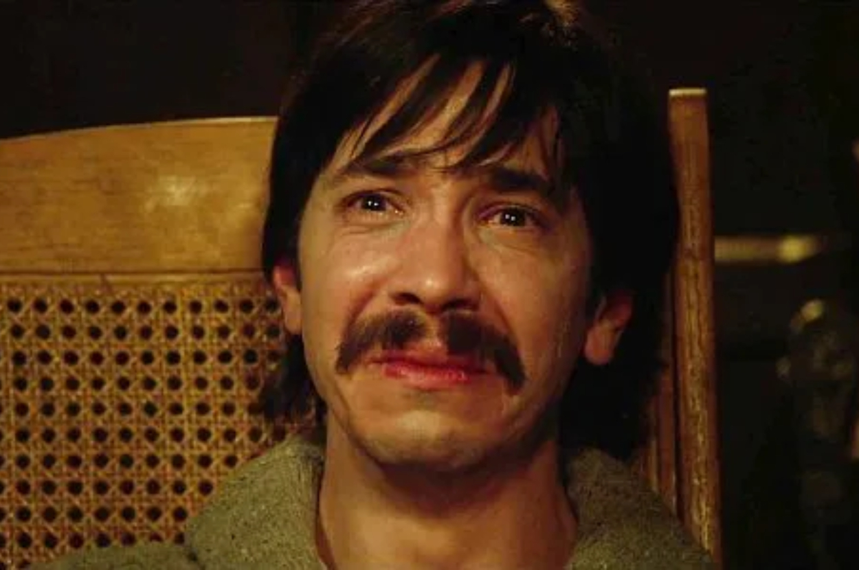 Man with mustache looks emotional, sitting in a chair. Appears to be a still from a TV show or movie