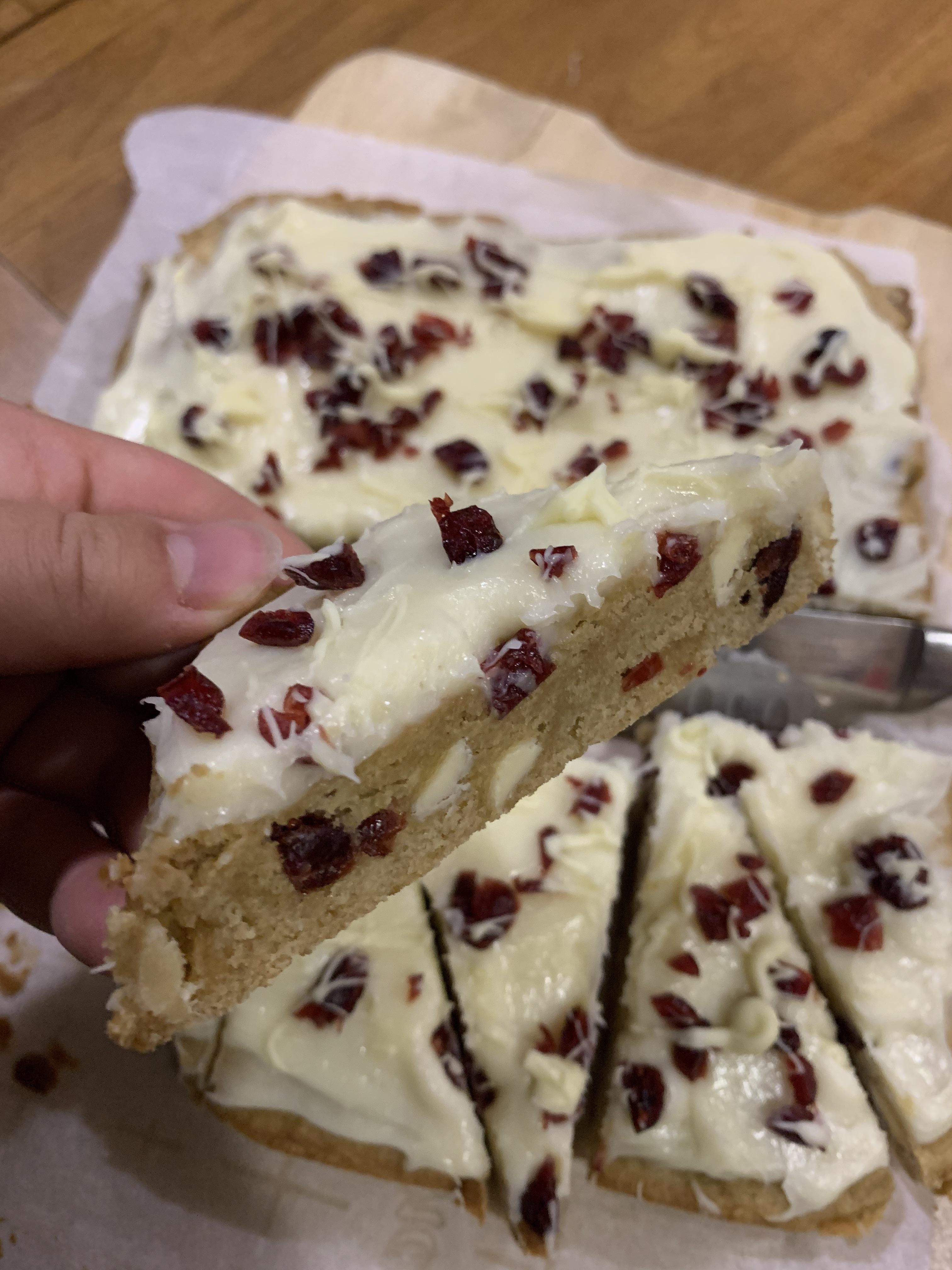 Hand holding a slice of cake topped with cream and cranberries, on a wooden table