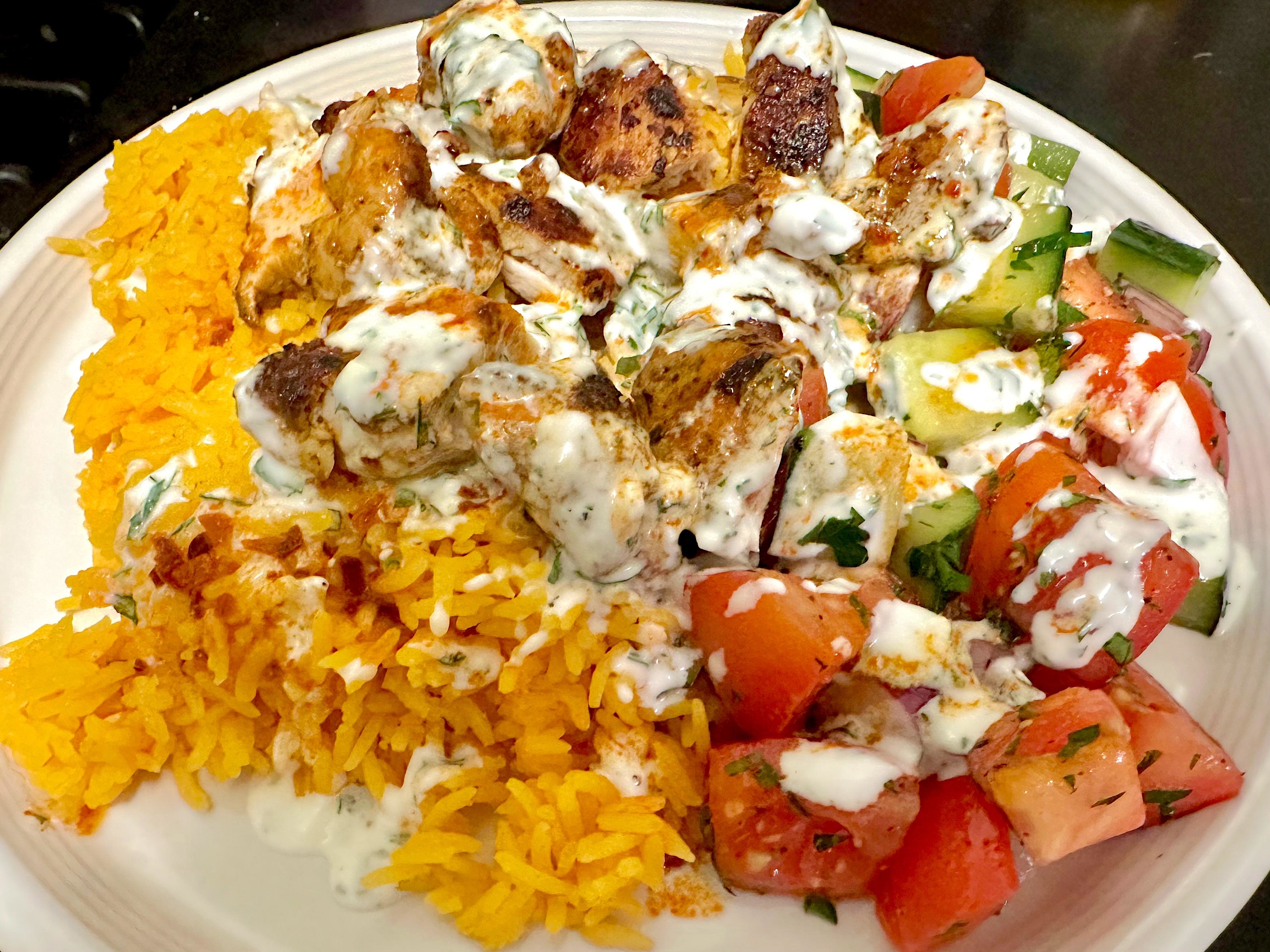 A plate of chicken kabobs with rice, chopped vegetables, and a creamy sauce