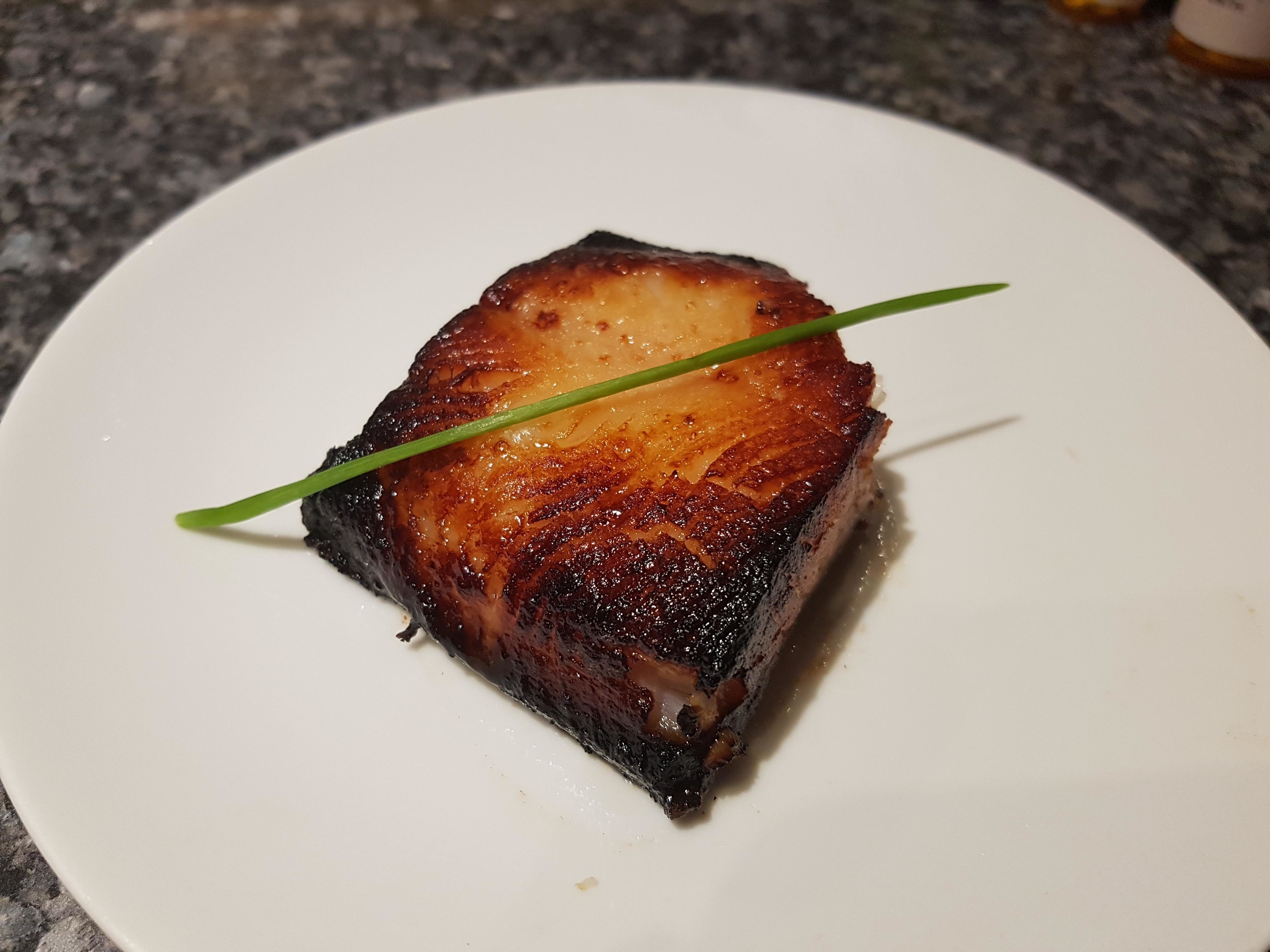 Seared pork belly slice on a white plate garnished with a green onion