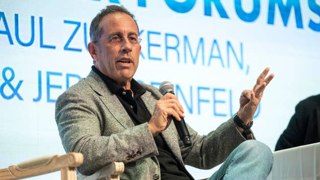 Jerry Seinfeld speaks at an event, seated, wearing a jacket and jeans, gesturing with his hand