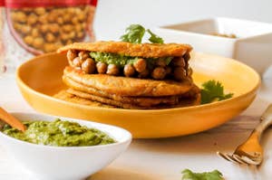 A stack of roti with chickpeas on top, next to a packet of A Dozen Cousins beans and a bowl of green sauce