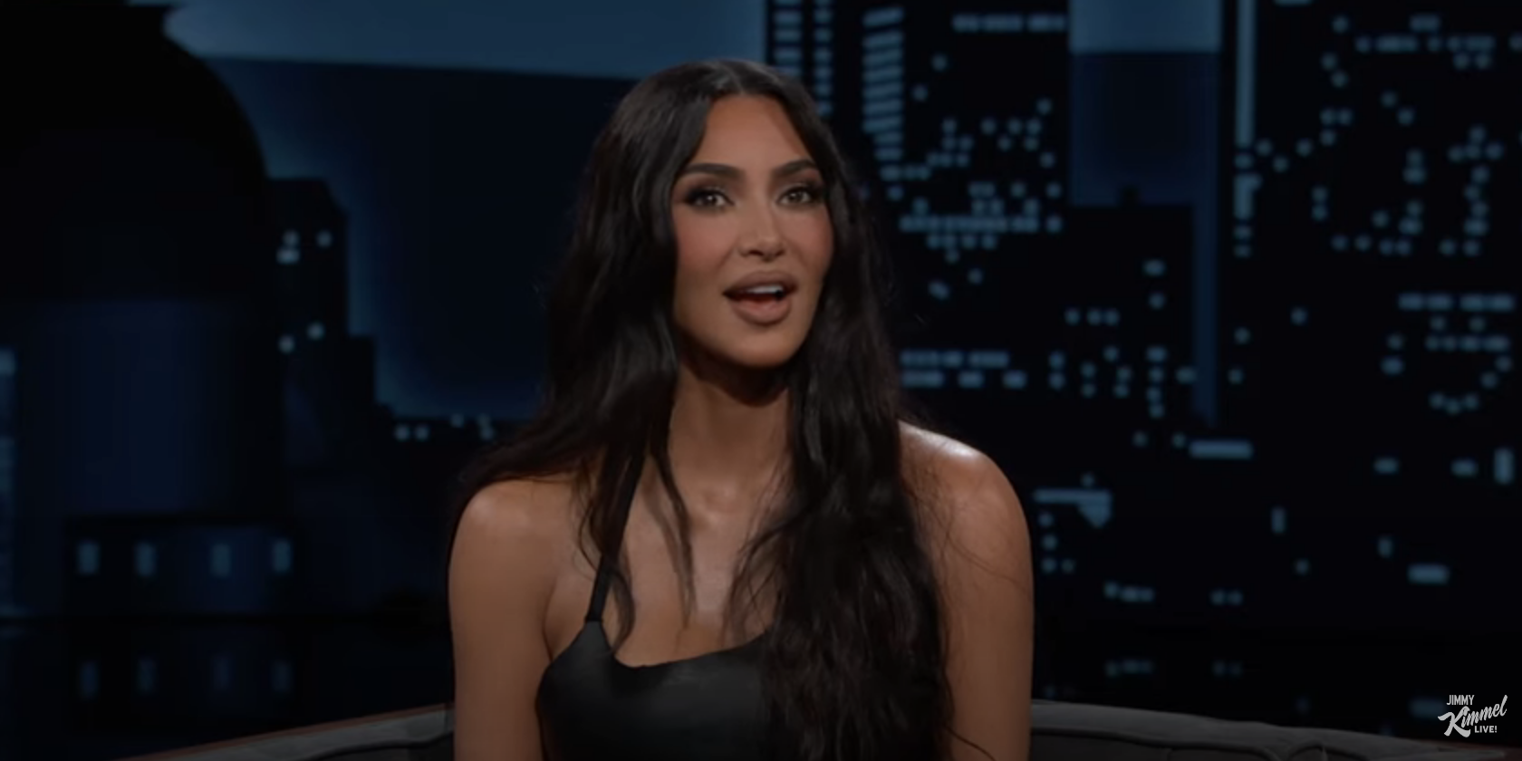 Kim Kardashian in a talk show setting, wearing a black top with a plunging neckline