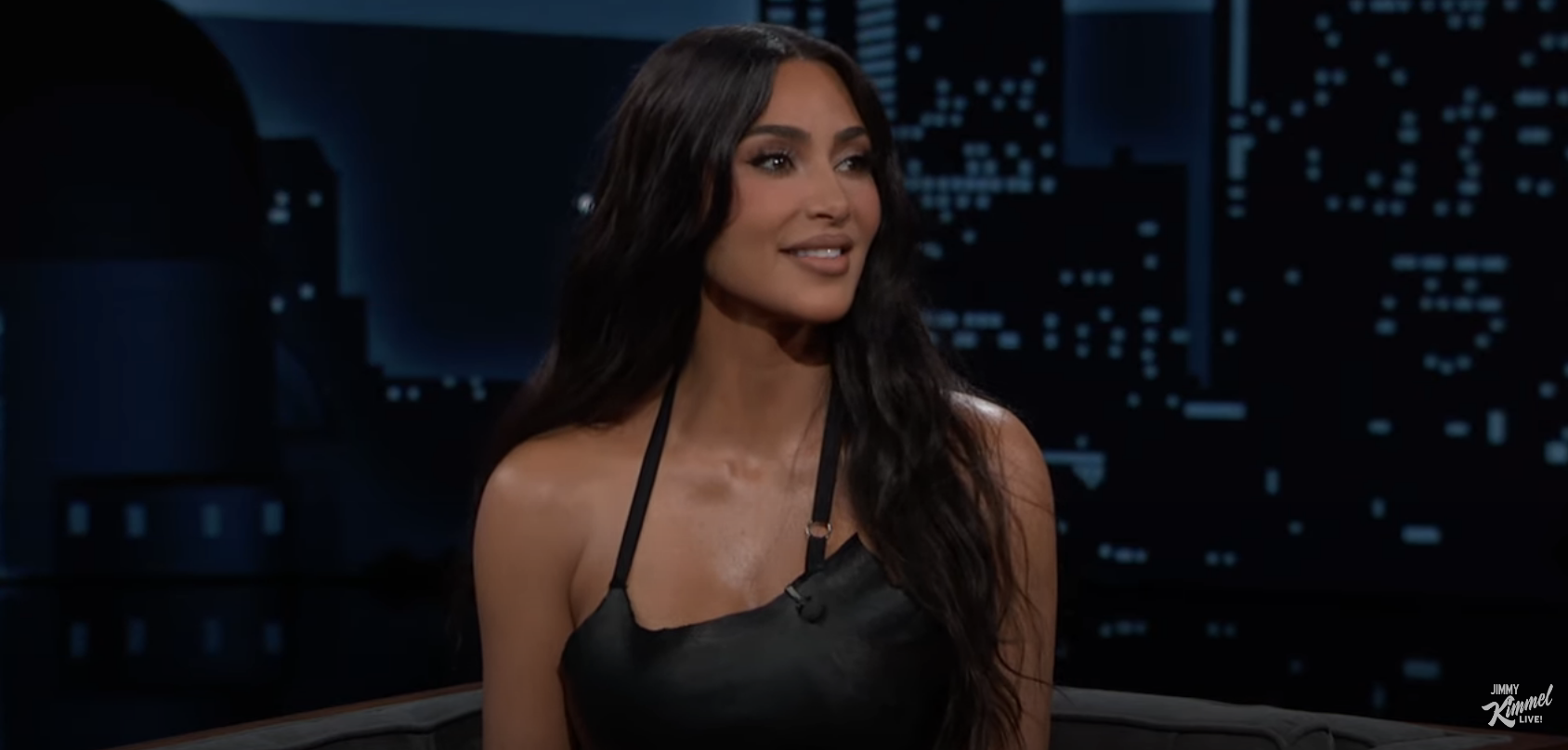 Kim Kardashian in a black top with a cityscape backdrop on a talk show set