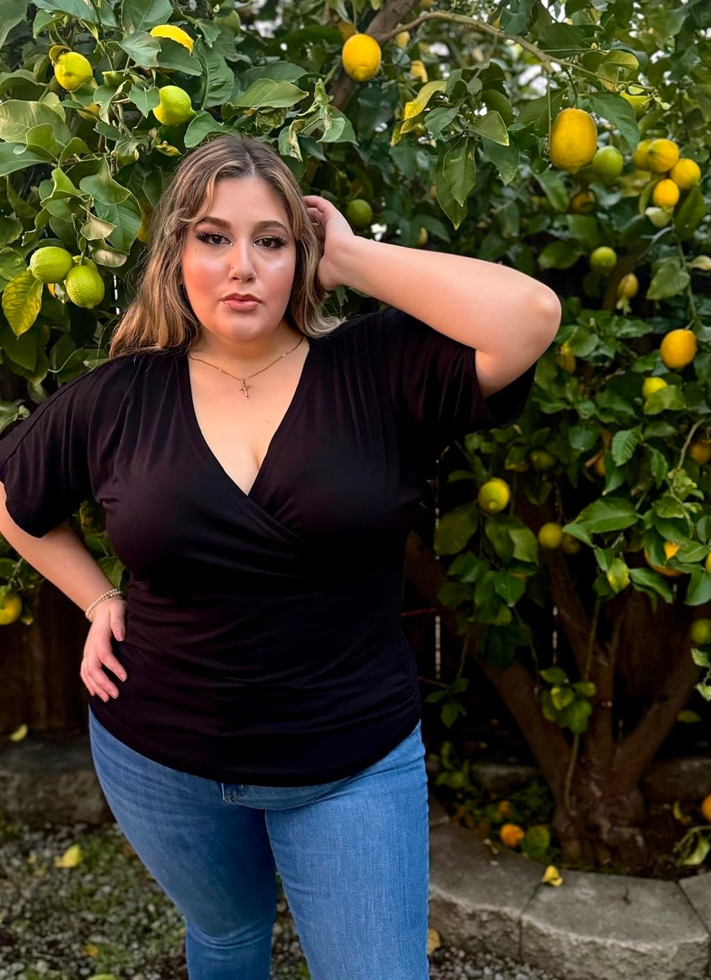 reviewer in a dark V-neck top and jeans poses with hand in hair by a lemon tree
