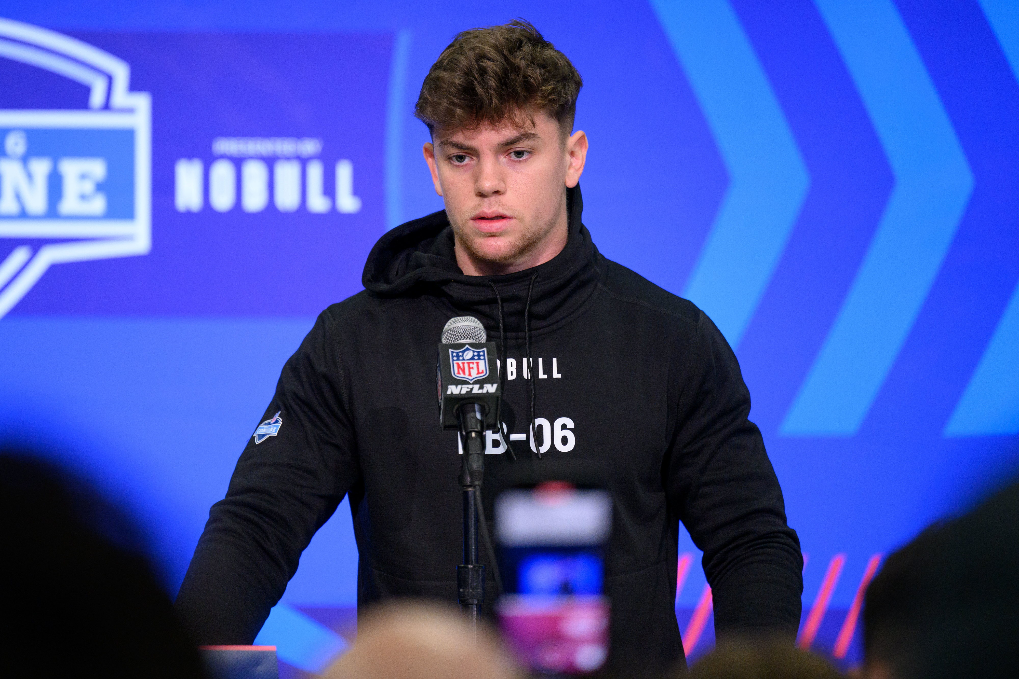 Person speaking at a podium with NFL and sponsor logos, wearing a sports hoodie with the text &quot;LB-06&quot;