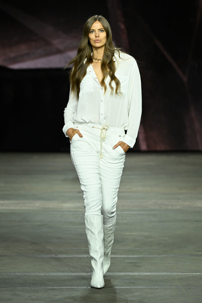Robyn walking on runway in light shirt, pants, and boots, fashion show setting