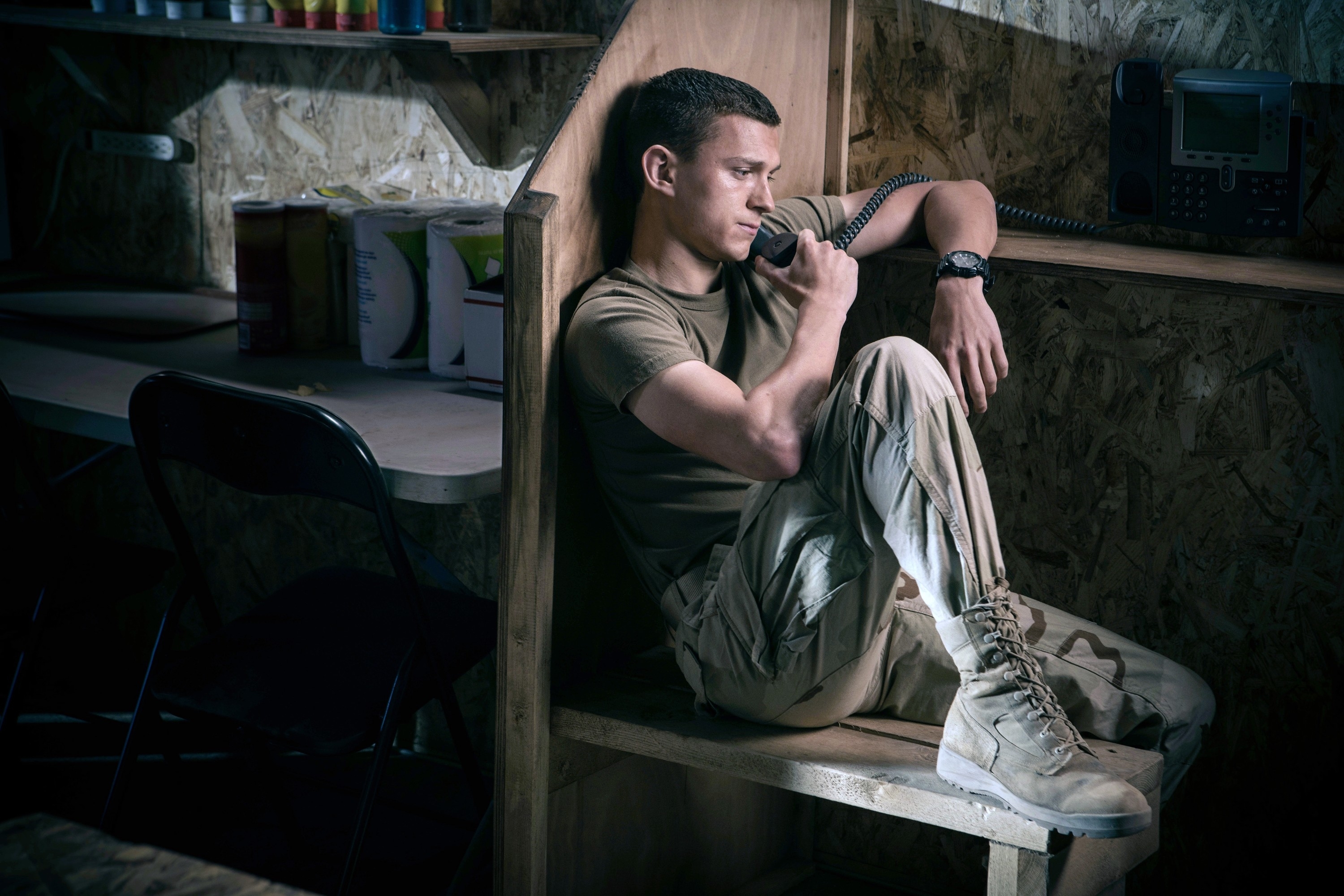 in a scene, Tom in military outfit sitting, talking on a corded phone, in a dimly lit wooden booth