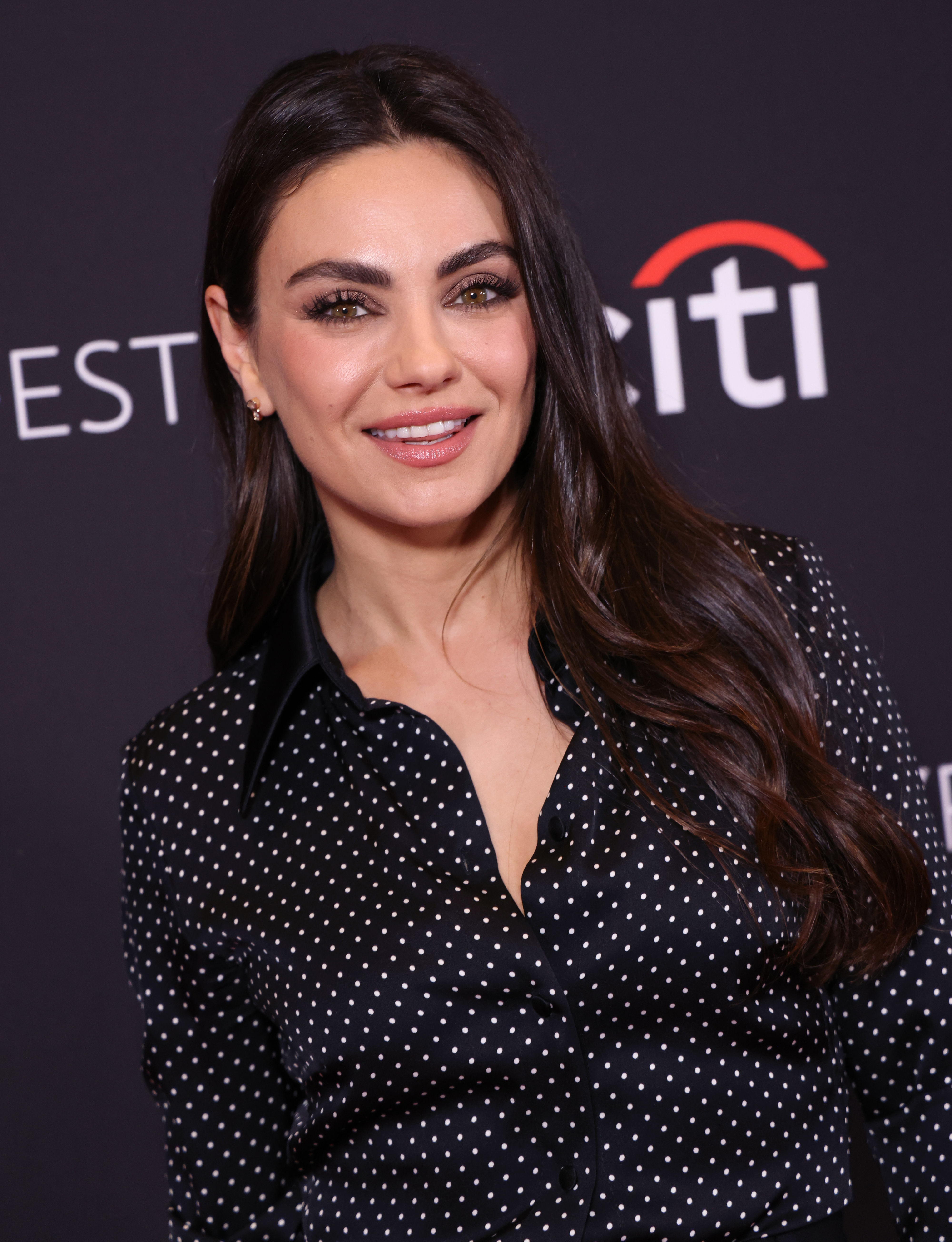 Mila Kunis poses with a smile and is wearing a polka-dotted top at a promotional event