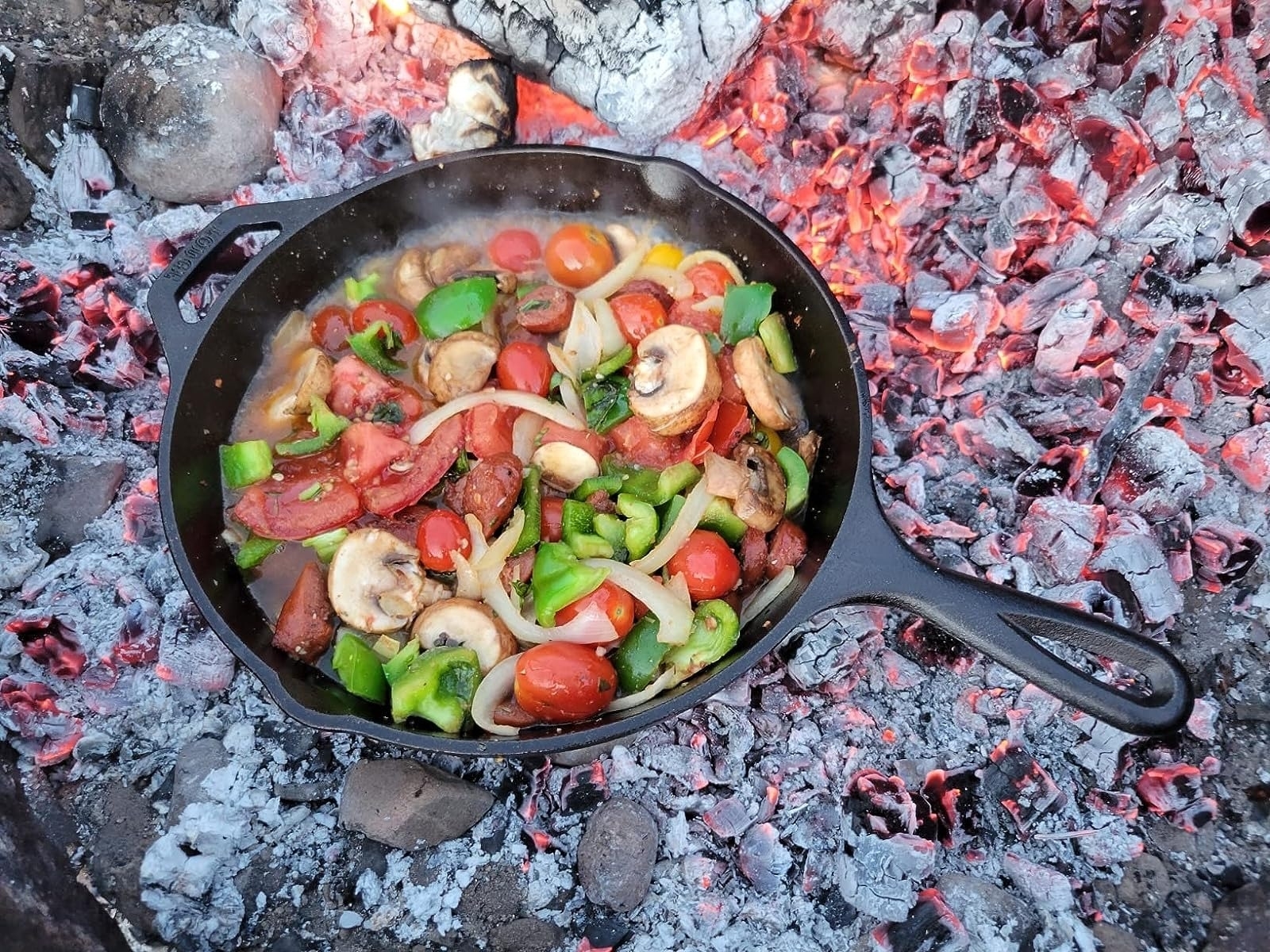 Cast iron skillet with mixed vegetables cooking over a bed of hot coals