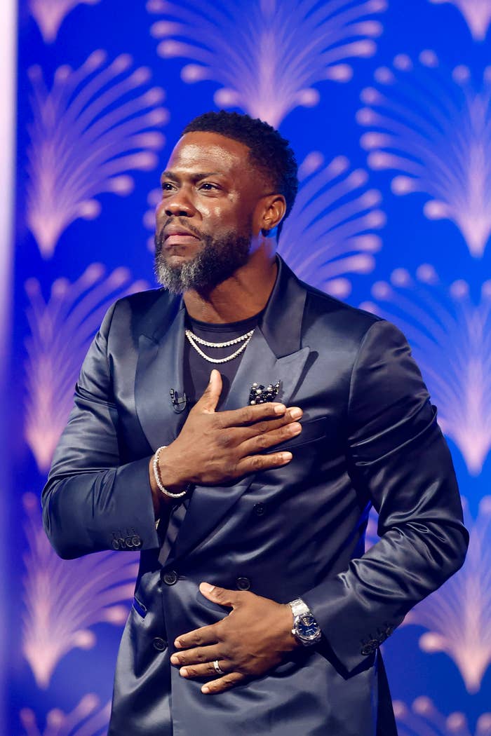 Kevin Hart in a stylish suit, standing with one hand over his heart and a serious expression