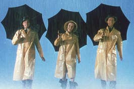 Three people in raincoats and hats holding umbrellas, appearing to dance